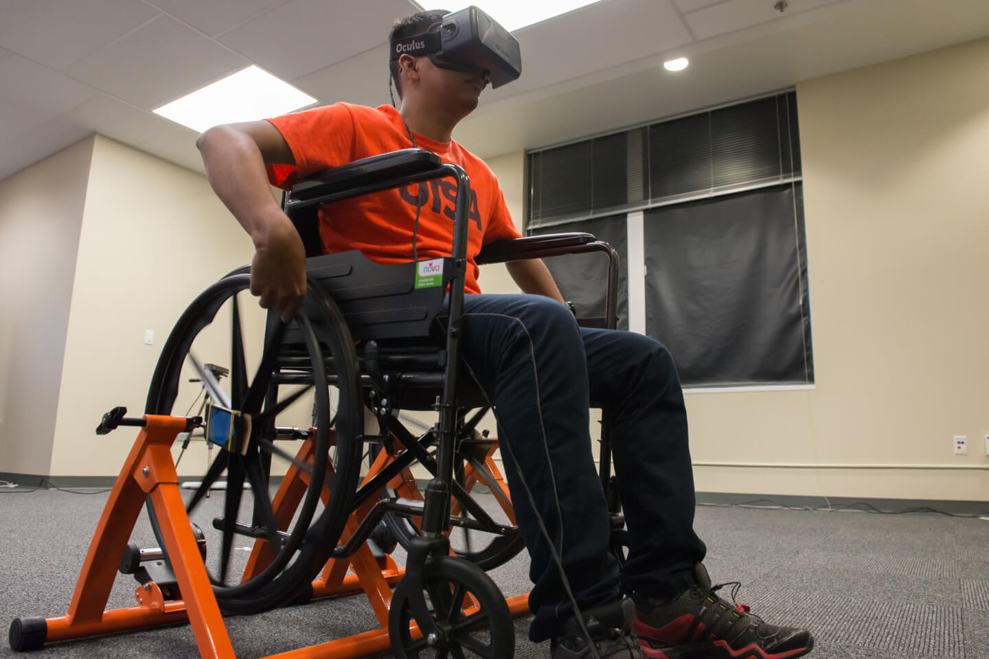 A man in an orange shirt is seated in a wheelchair wearing Oculus googles